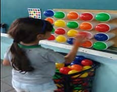 Uma menina, de costas, encaixa bolas coloridas num painel de furos, seguindo a sequência de cores da tabela ao lado.