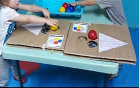 Duas crianças pequenas encaixam bolas coloridas em furos de um tabuleiro de papelão, seguindo os modelos de cores ao lado.