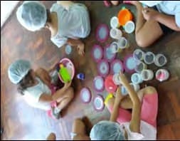 Crianças de touca, sentadas no chão, em volta de copos e potes de plástico de vários tamanhos.