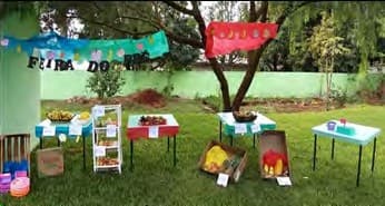 Feira montada no gramado da escola: mesas com frutas, legumes e preços, sob bandeirolas e uma faixa.