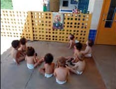 Bebês sentados em roda no pátio, diante de um muro vazado amarelo.