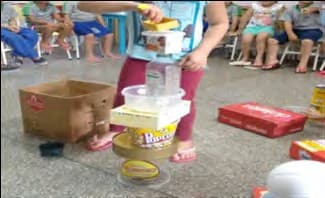 Uma criança empilha potes, latas e caixas de embalagens numa torre, no meio da roda de colegas sentados.
