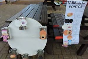 Num pátio, uma mesa redonda com latas enfeitadas de bichos e, ao lado, um cartaz com a tabela de pontos.