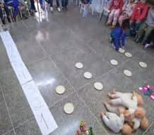 Gráfico no chão da sala, com pratinhos de papel enfileirados e brinquedos no início das colunas, observado por crianças sentadas.