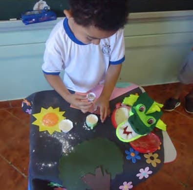 Um menino de uniforme, de pé diante de uma mesa forrada com tecido escuro e enfeites de feltro, compara potes e copos de tamanhos diferentes.