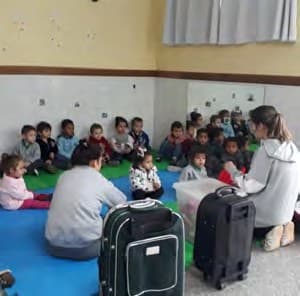 Crianças sentadas sobre tapetes, diante de duas professoras e de duas malas de viagem.