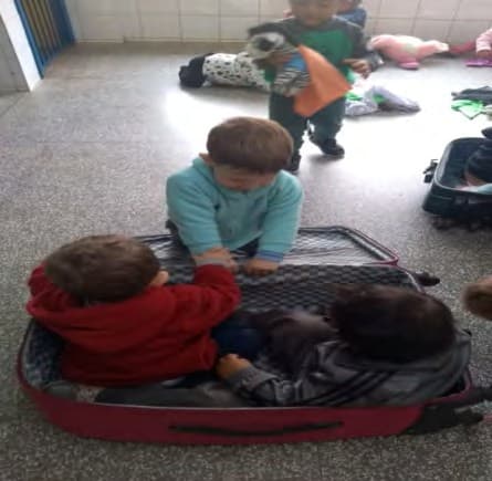 Três crianças dentro de uma mala de viagem aberta no chão da sala; outra observa ao fundo.