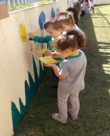 Crianças desenhando com giz num painel preso ao muro, ao ar livre, sobre a grama.