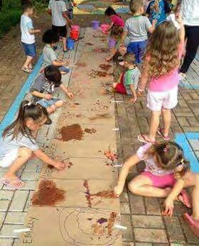 Crianças pintando com terra sobre uma faixa de papel estendida no chão do pátio.