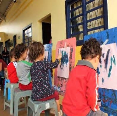 Crianças sentadas diante de cavaletes, pintando folhas presas a eles, na varanda.
