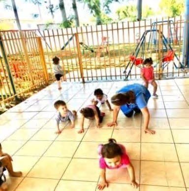 Uma professora e crianças engatinhando no piso de um pátio cercado.
