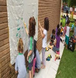 Crianças pintando um grande papel preso a um muro de tijolos, ao ar livre.