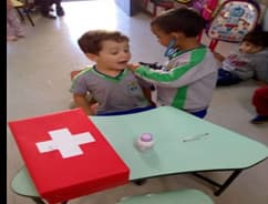 Dois meninos brincam de médico e paciente diante de uma maleta com uma cruz