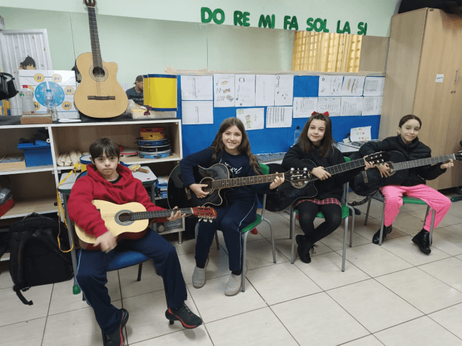 Crianças tocando violão em sala de aula, sob mural com a escala musical Dó-Ré-Mi.