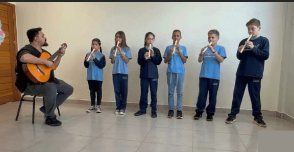 Crianças tocando flauta doce em fila, acompanhadas por um professor ao violão.