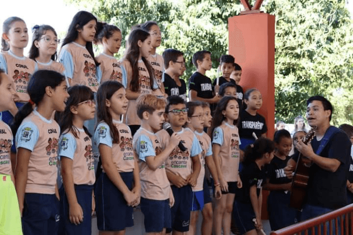 Coral infantil cantando ao ar livre, acompanhado por um professor ao violão.