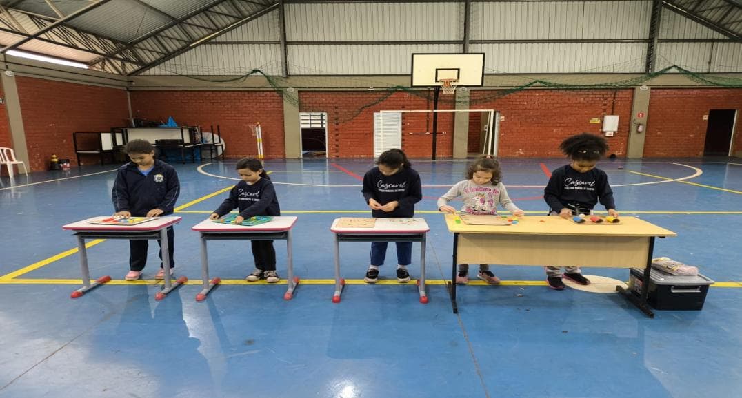 Crianças em fila jogando diferentes jogos de tabuleiro sobre carteiras no ginásio.